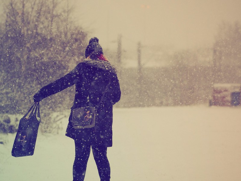 Shot from behind a woman as she walks through the snow as it falls. She is weating a winter coat and bobble hat, a Gorjuss bag on her shoulder and  her left arm is extended out and holding a Gorjust tote style bag in her hand.