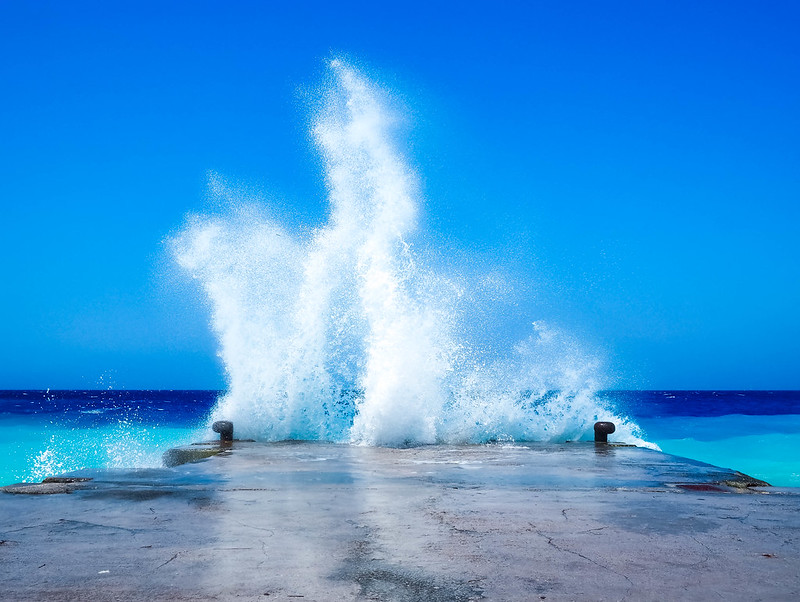 View of waves crashing into a concrete pier on the island of Rhodos, with a backdrop of the ocean. The sky is a light blue, the water at the horizon is a deep blue that gradually gradients through lighter blues and cyans. The view of the pier is straight on and waves reach over 10ft in height, fizzing and splashing.